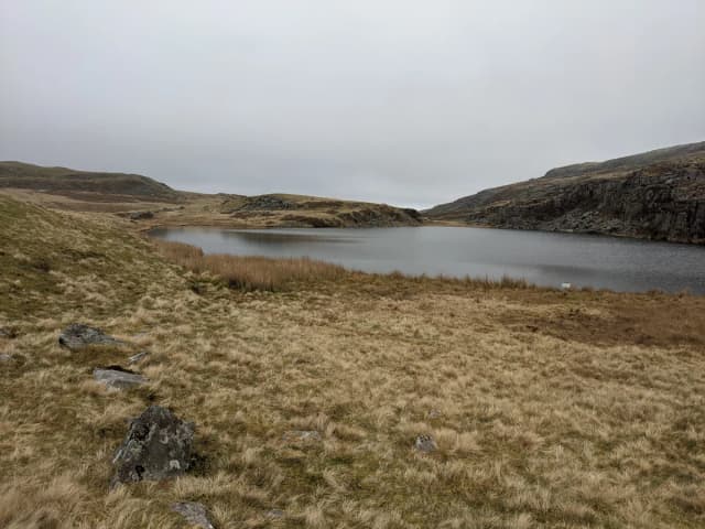 A grey, still upland lake bordered by tussocky moorland grass and a low rocky outcrop, under an overcast sky.