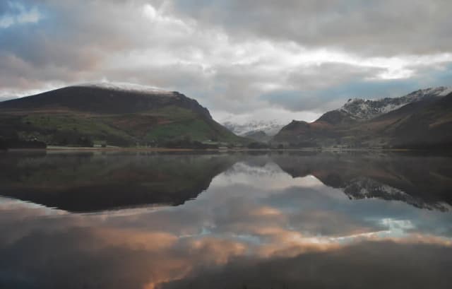Calm mirror-still Llyn Nantlle reflecting snow-capped Snowdonia mountains and a cloudy pink-tinged sunrise sky.