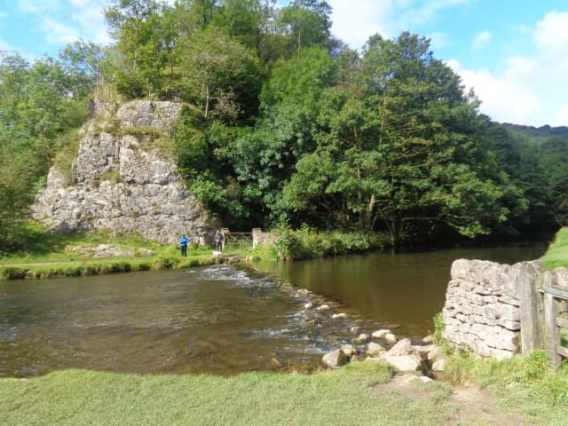 Shallow limestone river with stepping stones and a dry-stone wall, backed by a wooded limestone crag at Dovedale, Derbyshire.