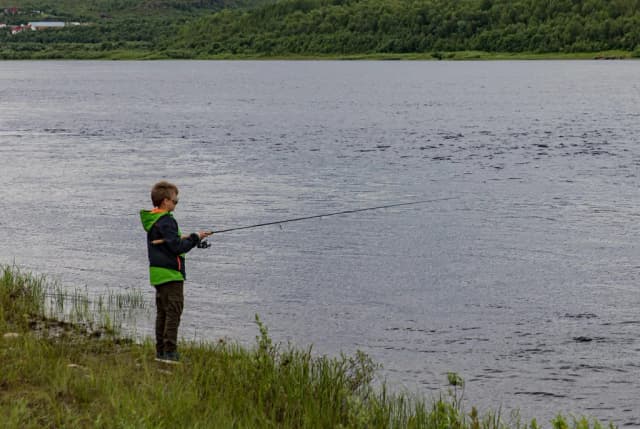 A boy fishing from the grassy bank of a wide river with forested far shore.