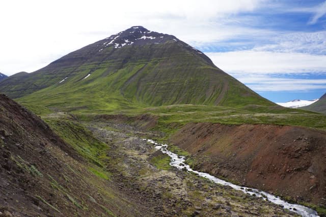 A small glacial river winding through a red volcanic gully beneath a green mountain in Iceland.