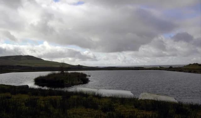 Wide view of the Glenquicken Fishery trout loch under a cloudy sky, with a small island and moorland hills beyond