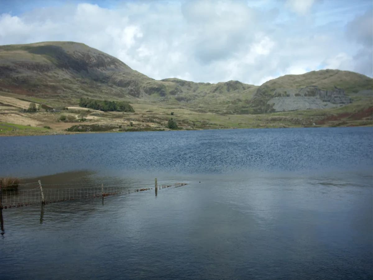 Still blue water of Llyn Cwmystradllyn with a submerged boundary fence, green hills, and an old slate quarry scar on the far hillside under a cloudy sky.