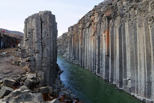 The glacial river Jökla flowing through the dramatic basalt column canyon of Studlagil in eastern Iceland, with hikers visible for scale on the canyon rim.