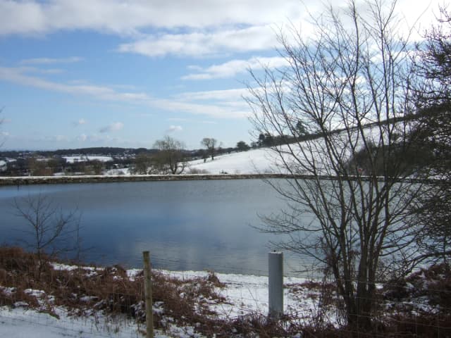 A small reservoir lake with an embankment dam, surrounded by snow-covered fields and bare trees, near Cilcain in the Clwydian Range.