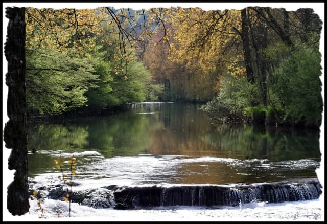 A small clear river flowing over a low weir, lined with autumn trees, in the Cusancin valley, France.