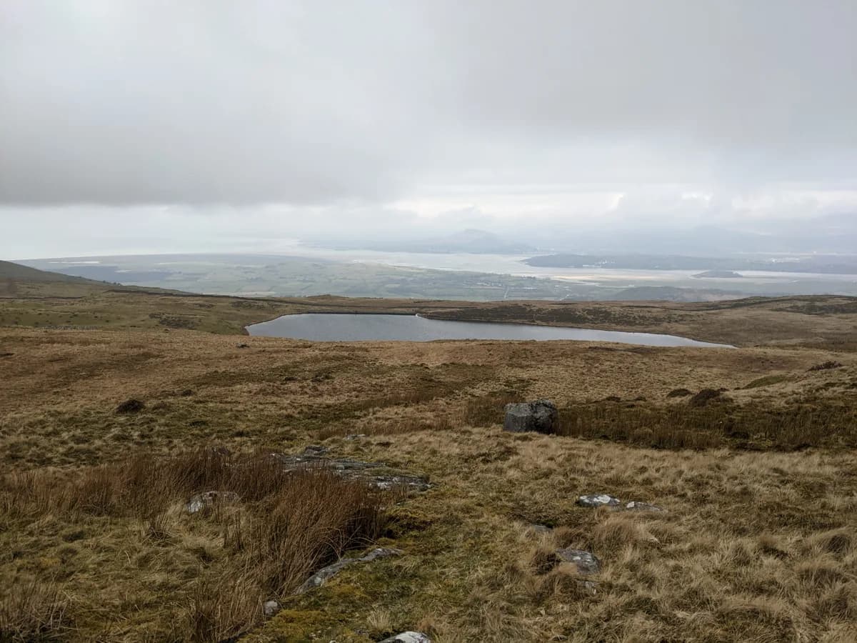 A small elongated moorland lake seen from a heather-covered hillside, with a flat coastal plain and misty hills in the far distance.