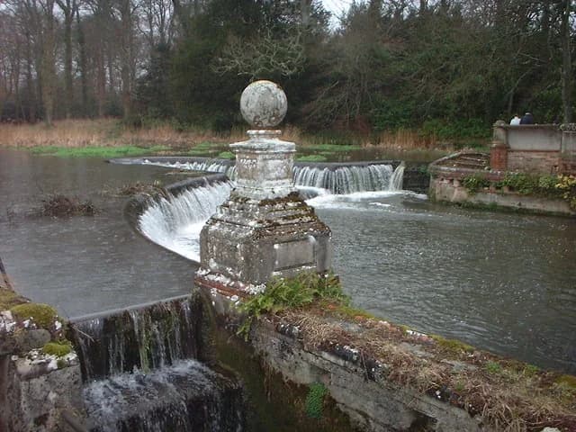 A weir on the River Piddle chalk stream near Trigon Farm, Dorset, with clear water flowing over the structure surrounded by rural countryside.