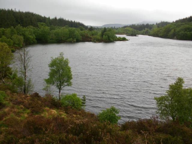 Wide view across a forest-ringed lake with wooded hills in the background, rain clouds overhead, near Betws-y-Coed.