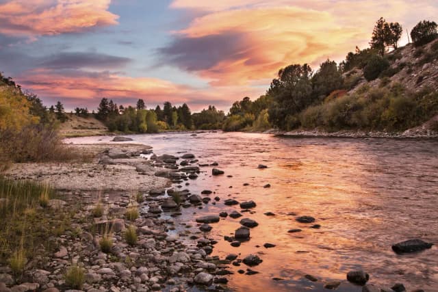 The Arkansas River flowing through a rocky streambed at sunset in Browns Canyon National Monument, Colorado, with pink and orange clouds reflected on the water.