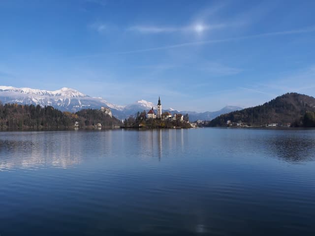 Lake Bled in autumn colours with calm reflective water, the island church mid-lake and forested slopes.