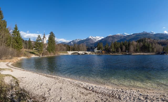 Panoramic view of Lake Bohinj from the shoreline near Stara Fužina, calm water, forested slopes and peaks.