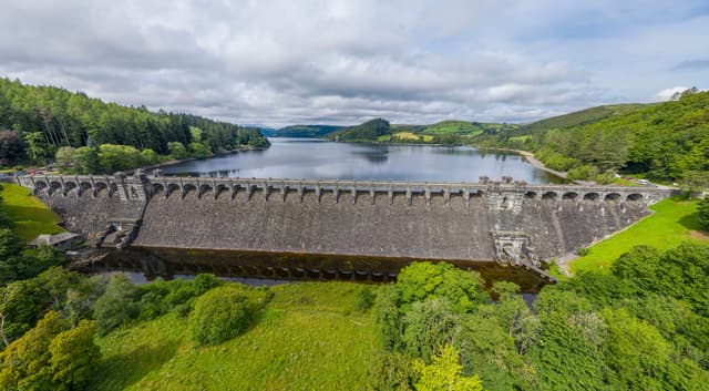 The stone dam wall of Lake Vyrnwy with the reservoir impounded behind it and wooded hillsides on the far bank.