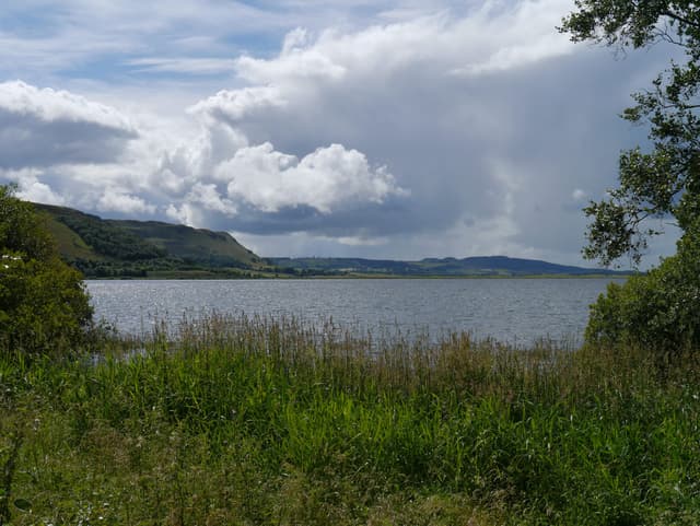 Broad view of Loch Leven from the south shore looking west, loch water filling the middle distance with farmland and hills beyond.
