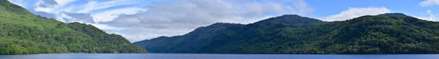 Ultra-wide stitched panorama of Loch Lomond showing the full breadth of open water, wooded islands and surrounding hills.