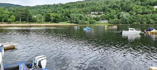 Panoramic view of Loch Tay at Kenmore showing the loch mouth, wooded shoreline and hills beyond under blue sky.