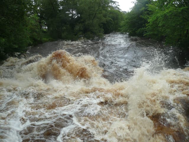 The River Ribble in full spate with brown peaty flood water over boulders near Settle.