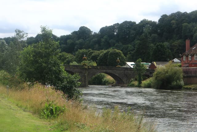 A tree-lined riverside footpath along the River Usk at the town of Usk, the broad river in view.