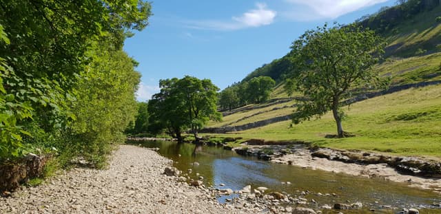 A wide panoramic view of the River Wharfe flowing through Kettlewell in upper Wharfedale below fell-flanked valley sides.