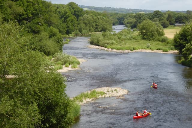The River Wye at Hay-on-Wye from the bank, broad open water with wooded hills behind, in summer.